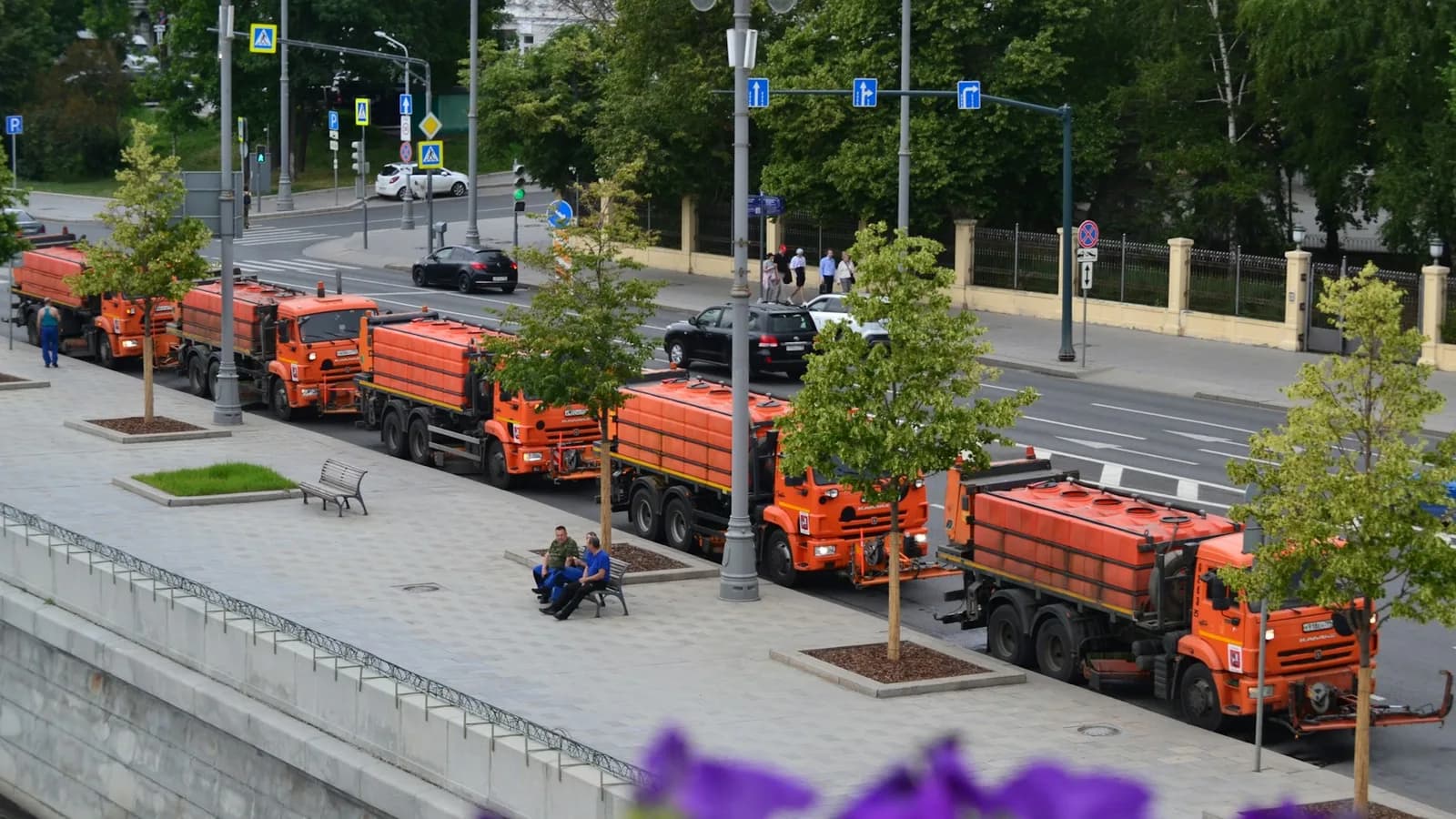 A row of orange street-cleaning trucks parked along a tree-lined city avenue